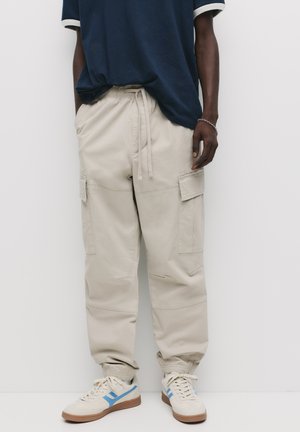 Pantaloni cargo bej cu manșete elastice și talie cu șnur. Prezintă multiple buzunare și o croială lejeră. Purtați cu adidași cu accente albastre.