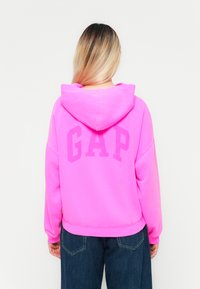 Rosa hettegenser med en stor "GAP" logo på baksiden. Laget av mykt materiale, har snøring på hetten, og har ribbestrikkede mansjetter og hem.