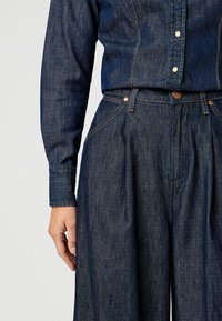 Henkilö, joka on pukeutunut tummansiniseen farkkupaitaan, joka on tuotu vyötärölle korkeisiin, leveisiin farkkuihin, joissa on metalliset napit ja ompelu yksityiskohtia.