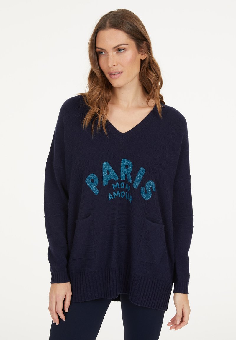 Navy blue oversized sweater with a V-neck, featuring a glittery "PARIS MON AMOUR" graphic and two front pockets. Ribbed hem and cuffs.