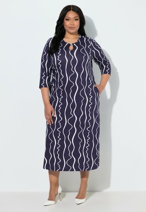 Donna che indossa un abito midi blu navy con righe verticali ondulate bianche, maniche tre quarti, scollo a chiave e tacchi alti bianchi.
