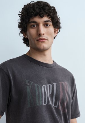 Νέος άνδρας με σγουρά σκούρα μαλλιά, φορώντας σκουλαρίκια καρφιά και ξεθωριασμένο μαύρο T-shirt με πολύχρωμο λογότυπο "The Kooples".