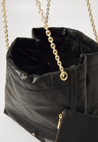 Sac seau en cuir matelassé noir avec une bandoulière en chaîne dorée et une poche zippée. Il présente une texture douce et un design intérieur compact.