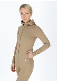 Beige Zip-Hoodie mit hohem Kragen, gerippter Textur und langen Ärmeln. Verfügt über seitliche Taschen und ein kleines Logo am unteren Ende.