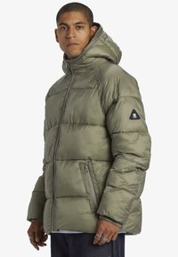 Olivgrön puffjacka med huva, med kviltad design, framzipper och en logotyp patch på ärmen. Två sidofickor synliga.