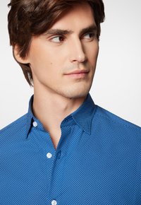 Niebieska koszula z guzikami, ozdobiona subtelnym białym wzorem w kropki. Kołnierz jest szeroki, a materiał ma gładką fakturę.