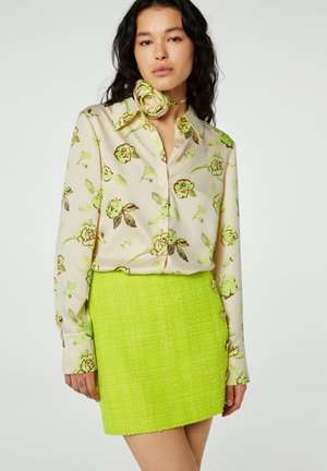 Vrouw draagt een crèmekleurige bloemenblouse met groene en bruine bloemen, gecombineerd met een fel limoengroene getextureerde minirok en een bloemenhalsdoek.