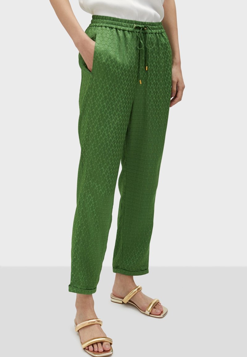 Person wearing green patterned drawstring pants with rolled cuffs and gold open-toe sandals, standing with one hand in pocket.