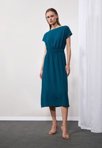 Teal midi šaty s krátkými rukávy, elastickým pasem a hladkou strukturou. Doplněné béžovými sandály na vysokém podpatku na neutrálním pozadí.