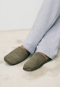 Brązowe teksturowane klapki z żebrowanym wzorem i solidną podeszwą, w połączeniu z szarymi spodniami dresowymi. Obuwie ma zamknięty przód i jest zakładane na stopę.