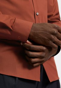 Rust-colored long-sleeve shirt with a smooth texture, button closure, and contrasting cuffs. Features subtle logo embroidery near the wrist.