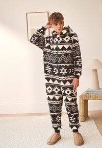 Tuta intera in pile nero e crema con motivi geometrici, zip frontale e cappuccio. Abbinata a pantofole slip-on color castagna. Tessuto strutturato.