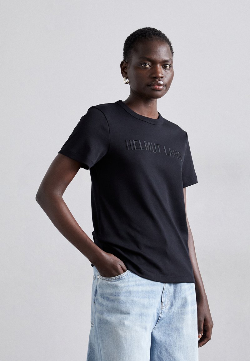 T-shirt de algodão preto com mangas curtas e gola redonda, apresentando o logótipo em relevo "HELMUT LANG" no peito, combinado com jeans de ganga claros.