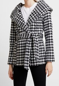 Cappotto a fantasia houndstooth bianco e nero con un colletto oversize, vita cinturata e maniche lunghe. Realizzato in tessuto testurizzato.