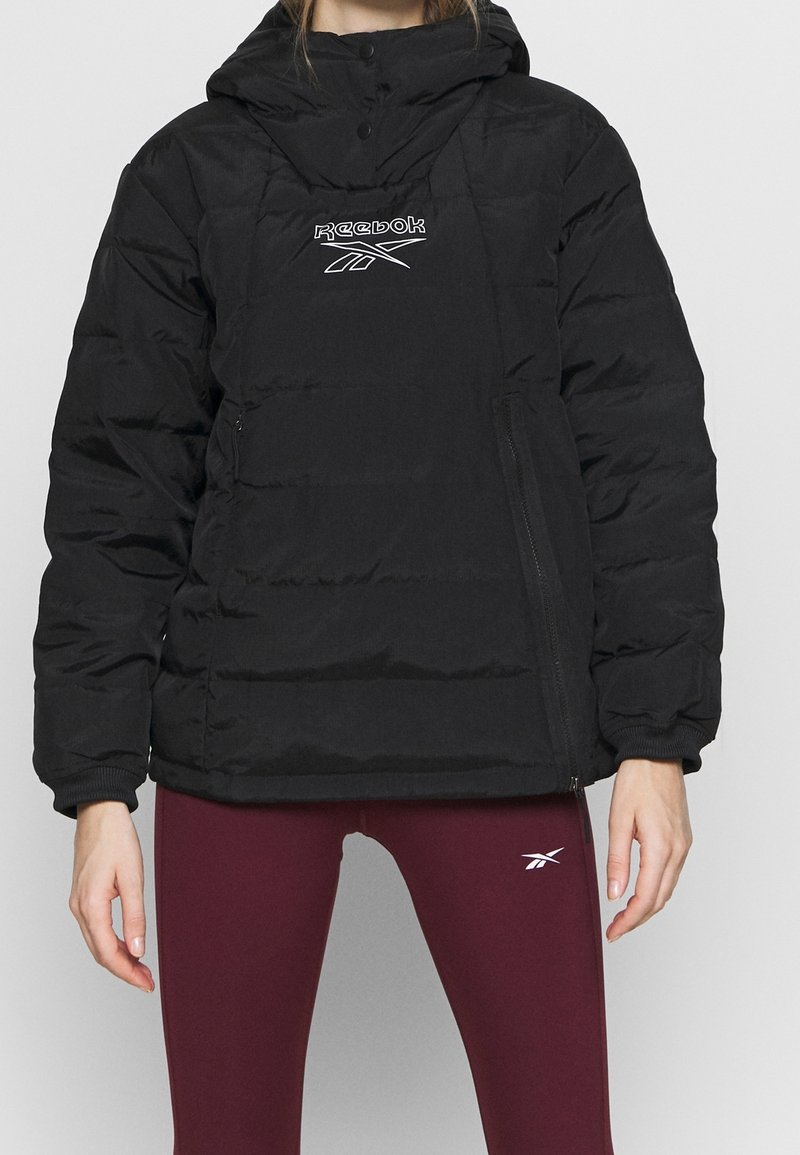 Personne portant une doudoune noire Reebok avec capuche et des leggings bordeaux avec un logo Reebok blanc sur la cuisse.