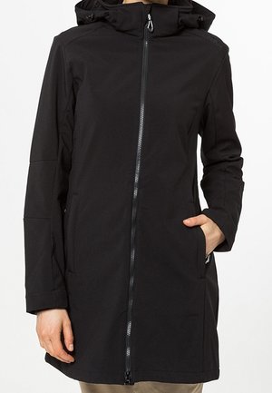 Personne portant un long manteau noir à capuche avec une fermeture éclair sur le devant, une main dans la poche, debout devant un fond clair uni.