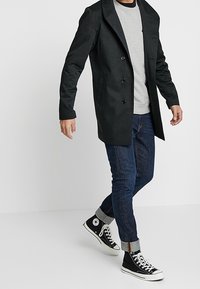 Cappotto nero doppiopetto con ampi risvolti, indossato sopra una felpa grigia. Abbinato a jeans in denim scuri e sneaker nere alte.
