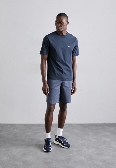 Camiseta de algodón azul marino con mangas cortas y cuello redondo, combinada con pantalones cortos grises. El modelo lleva zapatillas azul marino con suelas blancas y calcetines.