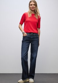 Mujer de pie con la mano en el bolsillo, vistiendo una camiseta roja de manga corta, vaqueros de pierna ancha azul oscuro con cinturón, pulseras de plata y zapatillas blancas.