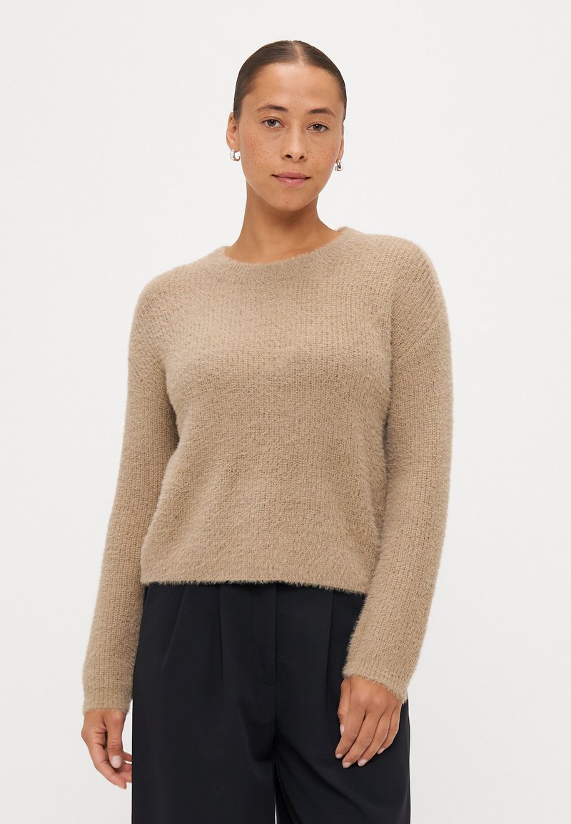 Beige kötött pulóver puha, texturált felülettel, kerek nyakkivágással és hosszú ujjakkal, sötét nadrággal párosítva.