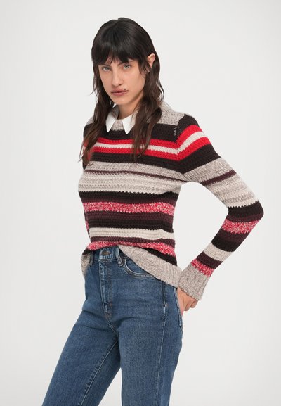 Hosszú sötét hajú nő, aki színes csíkos kötött pulóvert visel fehér inggel és kék farmerrel, egyik kezét hátul tartva áll.