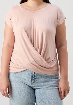 Frau trägt ein hellrosa kurzärmeliges Top mit verdrehtem Vorderteil, kombiniert mit hellblauen hoch taillierten Jeans, steht vor einem schlichten Hintergrund.