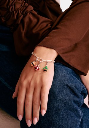 Hand mit silbernem Gliederarmband mit Anhängern in Form einer Biene, einer roten Teekanne und eines grünen Kaktus, ruht auf blauem Denim und braunem Ärmelsaum.