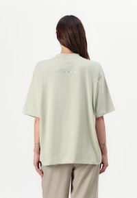 Hele roheline oversize t-särk, mis on valmistatud pehmest kangast, koos ümarate kaelustega ja seljateksti logoga, mis on valmistatud peegeldavast materjalist. Lühikesed varrukad.