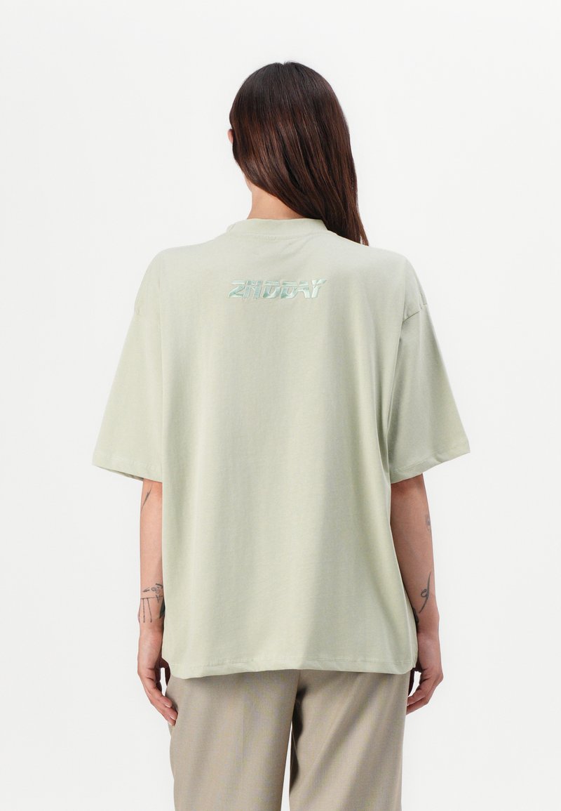 Hele roheline oversize t-särk, mis on valmistatud pehmest kangast, koos ümarate kaelustega ja seljateksti logoga, mis on valmistatud peegeldavast materjalist. Lühikesed varrukad.