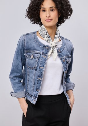 Femme aux cheveux bouclés portant une veste en jean bleu clair, une chemise blanche, un pantalon noir et une écharpe blanche à motifs nouée autour du cou.