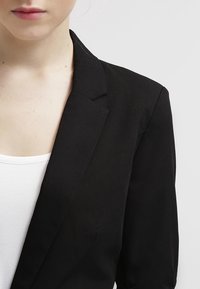 Czarny żakiet z wyraźnym kołnierzem i gładką teksturą. Noszony na białej bluzce, podkreślający dopasowany krój i minimalistyczny design.