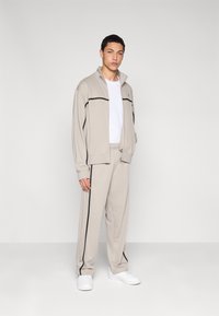 Μπεζ αθλητική φόρμα με ζακέτα με φερμουάρ και παντελόνια με ελαστική μέση. Μαύρες ρίγες στη ζακέτα και στα πόδια. Λευκό πουκάμισο και αθλητικά παπούτσια.