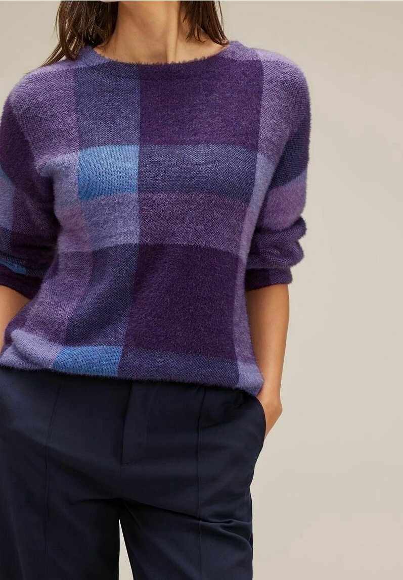 Femme portant un pull doux à carreaux violets et bleus avec les manches retroussées, associé à un pantalon bleu marine foncé, main dans la poche.