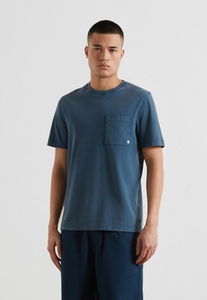 Jonge man die een gewassen blauw T-shirt met korte mouwen en borstzak draagt en een donkerblauwe broek, staande tegen een effen lichte achtergrond.