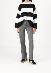 Personne portant un pull à larges rayures noires et blanches, un jean gris taille haute à jambes droites, et des baskets noires à rayures blanches.