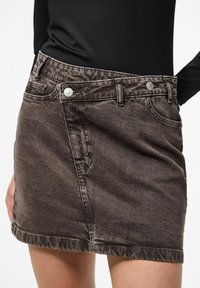 Frau trägt einen ausgewaschenen schwarzen Denim-Minirock mit zwei silbernen Knöpfen und Taschen, kombiniert mit einem schwarzen Langarmtop.