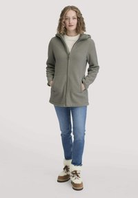 Langärmliger, mit Reißverschluss versehenen Fleece-Mantel in Olivgrün mit Kapuze, zwei Fronttaschen, kombiniert mit blauen Jeans und braunen, mit Fell gefütterten Stiefeln.