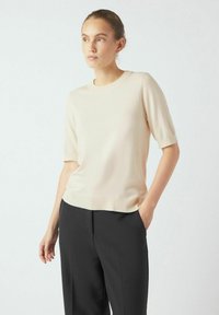 Top corto a maniche, girocollo, in morbido maglione beige, con orlo a costine, abbinato a pantaloni neri, presentato su uno sfondo neutro.