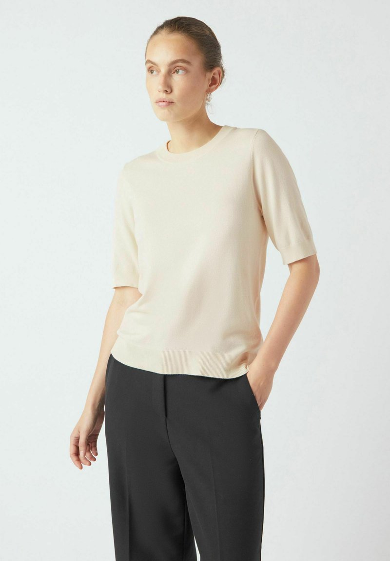 Top corto a maniche, girocollo, in morbido maglione beige, con orlo a costine, abbinato a pantaloni neri, presentato su uno sfondo neutro.