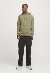Olive green hoodie with drawstring, front pocket, and white logo. Paired with black cargo pants featuring side pockets and a smooth texture.