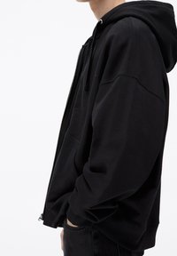 Personne portant un sweat à capuche noir zippé, les mains dans les poches, vue de profil sur un fond blanc uni.