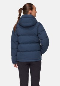 Kvinna med flätad hästsvans som bär en blå vadderad jacka med huva och svarta byxor, står med händerna i fickorna och vänder ryggen åt.