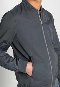 Chaqueta bomber gris hecha de tela suave, con cierre de cremallera en el frente, puños acanalados y bolsillos laterales. Diseño minimalista con cuello alto.