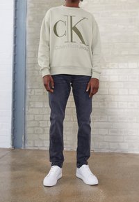Sudadera verde claro con un audaz logo "CK", combinada con vaqueros oscuros y zapatillas blancas. Tela suave con puños y dobladillo acanalados.
