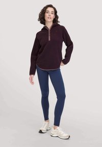Burgunderfarbener Fleece-Pullover mit halbem Reißverschluss, langen Ärmeln und abgerundetem Saum, kombiniert mit marineblauen Leggings und hellen Turnschuhen.
