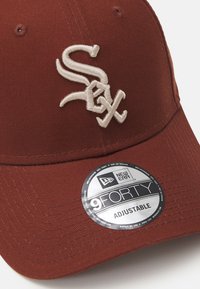 Casquette de baseball marron avec logo blanc brodé. Elle présente un design structuré, une sangle ajustable et une étiquette circulaire indiquant la taille.