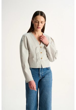 Kobieta o długich ciemnych włosach nosi koszulę w kratę z bufiastymi rękawami i drewnianymi guzikami oraz niebieskie, wysokie dżinsy na białym tle.