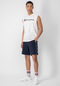 Biely tielko s kapucňou bez rukávov s logom "Champion", tmavomodré šortky, šedé ponožky s červenými a modrými pruhmi a biele športové topánky.