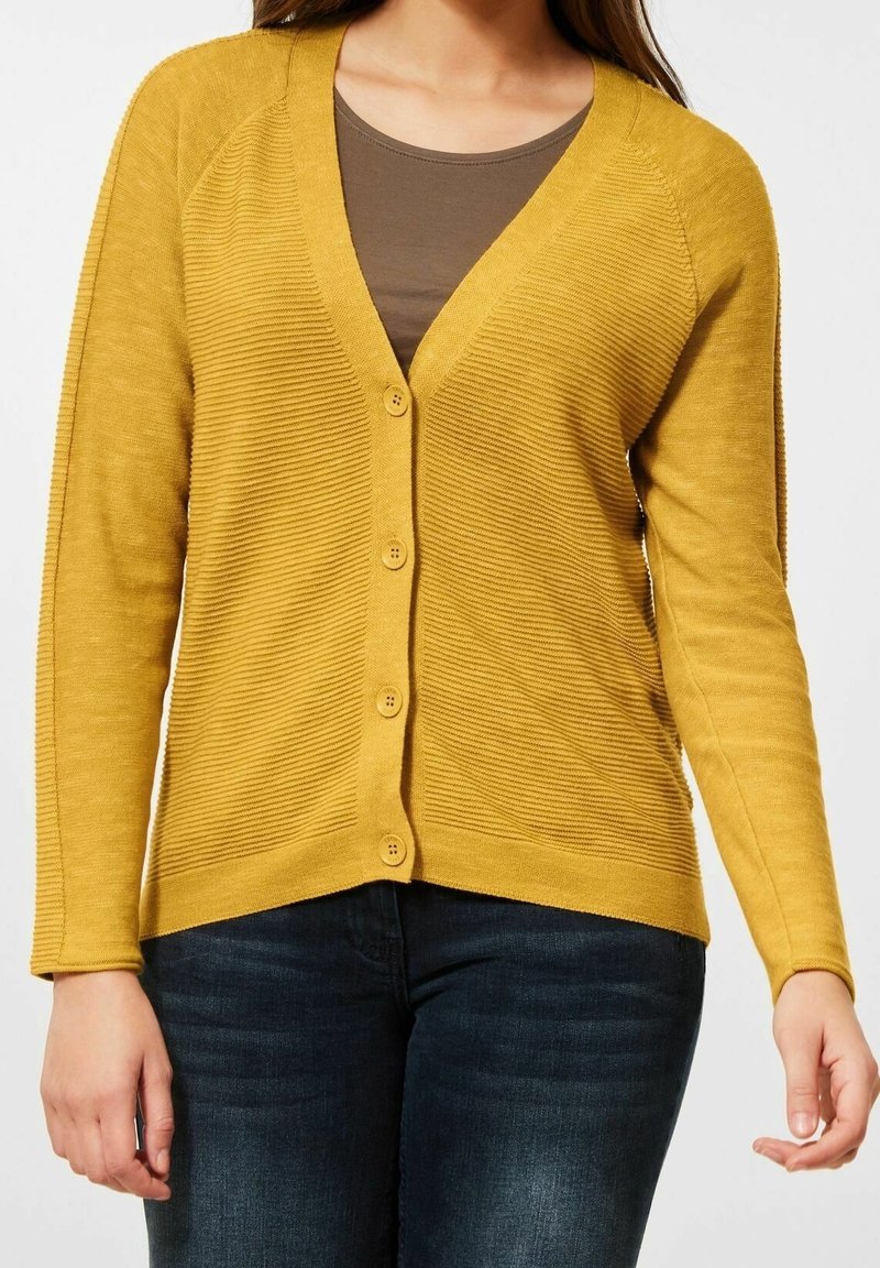 Senapsgul cardigan med ribbat mönster, V-ringning, knapplåsning och långa ärmar. Parat med en brun topp och mörka jeans.