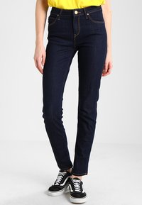 Hög midja mörkblå jeans i denim, utrustade med fem fickor, synlig sömnad och slim fit, kombinerade med svarta sneakers.